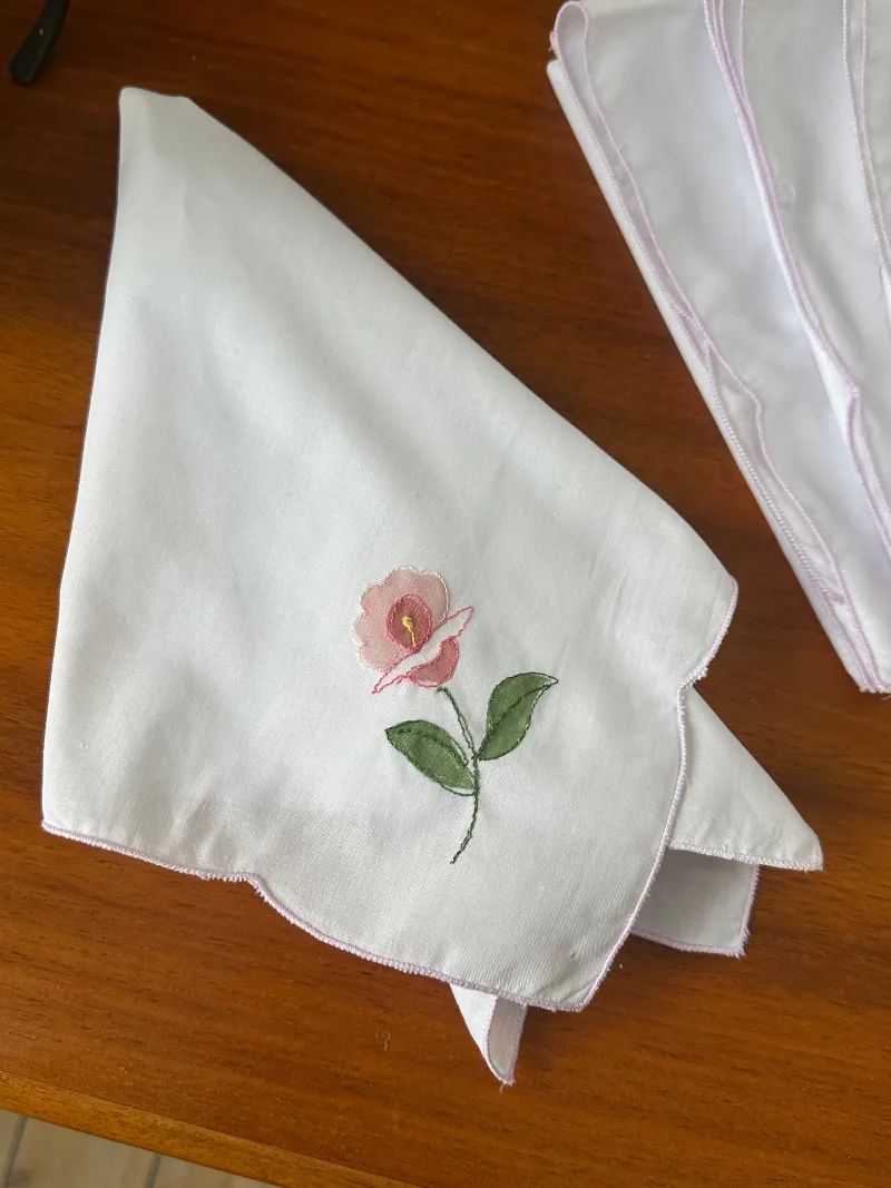 Tygservetter i vitt med rosa blomma och ljuslila broderad kant på ett träbord.