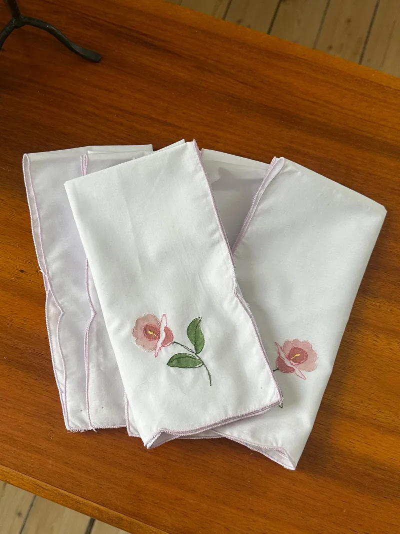 Sex vita tygservetter med ljuslila broderad kant och rosa blommor ligger på ett träbord.