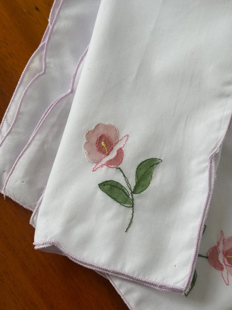 Sex stycken vita tygservetter med rosa broderad blomma och ljuslila broderad kant ligger på ett träbord.