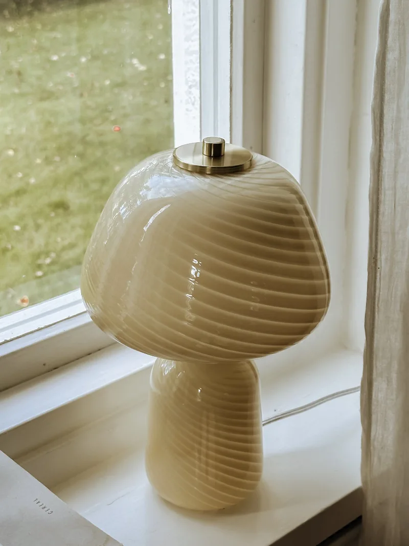 Bordslampa Bijou i bordeauxfärgat glas med spiralmönster och detaljer i mässing står på en vit fön利益bräda.