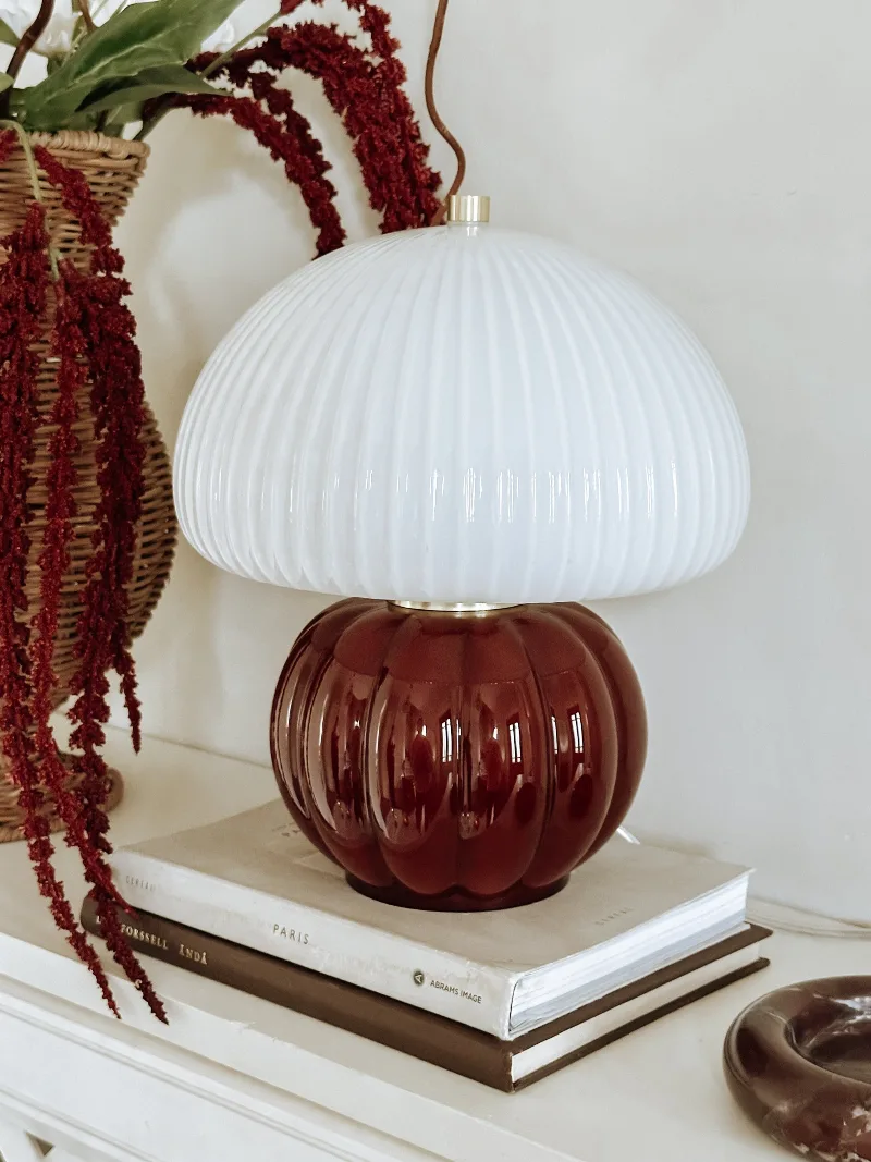 Bordslampa Margaux Bordeaux med bordeauxröd glasfot och rundad skärm i vitt räfflat glas på en vit byrå.